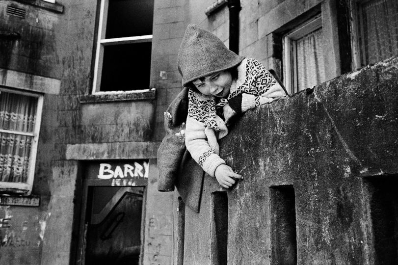 Child scaling a wall. Possilpark, Glasgow. 1986. Possilpark was one of the poorest in the city. It’s decline began when in 1967 the Saracen Foundry, which was the main employer in the area, closed down. During the 1980s Possilpark had become the hub of the city’s heroin trade. High levels of anti social behaviour and burglary made life stressful. People I met refused to leave their flats unoccupied even to go to the shops for fear that in their absence their flats would be emptied. © Mark Abrahams - Courtesy of Bluecoat Press